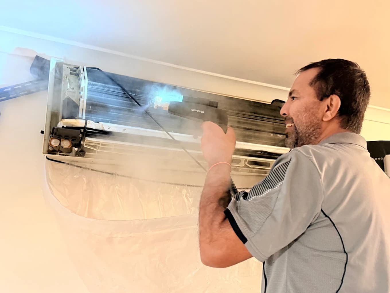 Technician steam-cleaning an aircon unit, coils exposed with visible steam
