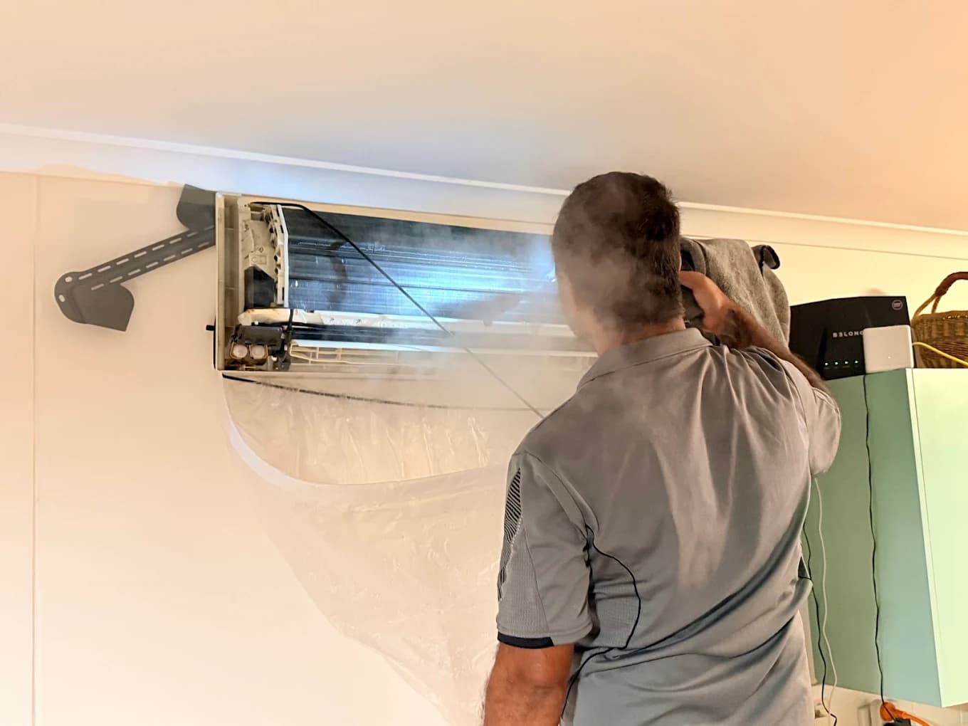 Technician steam-cleaning an aircon unit from behind, coils exposed with visible steam